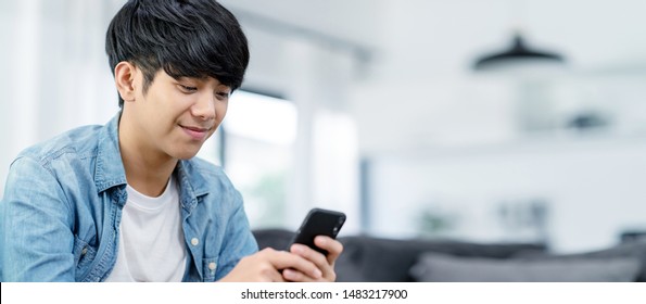 Panoramic or Banner of Happy asian teenager using smart phone and smiling on sofa living room at home. Asian man holding and using cellphone for searching data and social medie on internet.