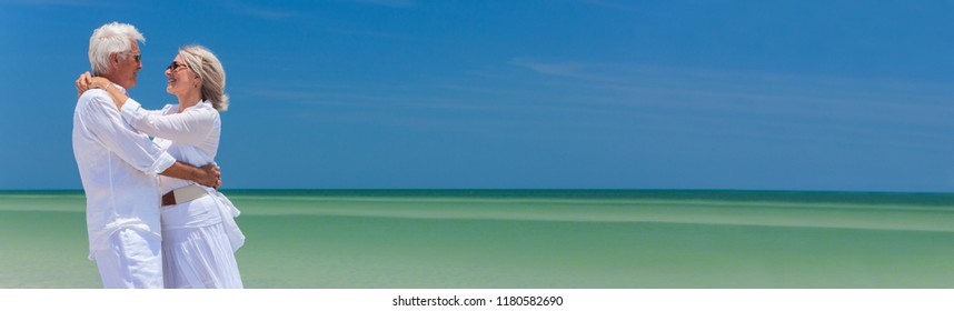 Panoramic banner image of a happy senior man and woman couple together embracing by sea on a deserted tropical beach with bright clear blue sky