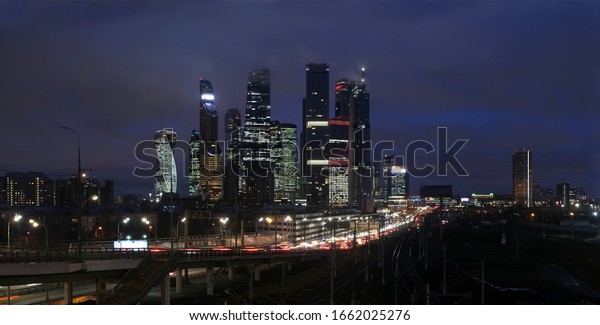 Panoramic city landscape with many\
railroad tracks going far, cars on the overpass and skyscrapers\
with towers of Moscow City with lighting windows in the\
evening