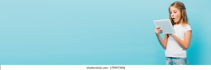 panoramic concept of surprised girl in white t-shirt using digital tablet isolated on blue