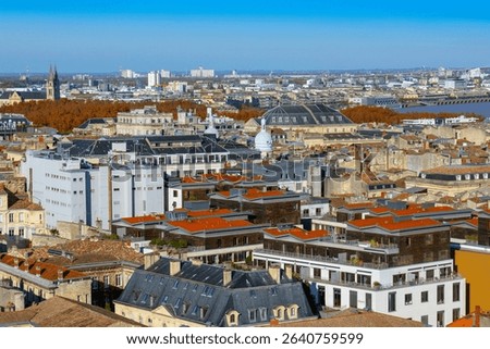 Panoramic elevated view of central and northeastern Bordeaux, showing dense old city rooftops, with a river, bridge, and hills with buildings in the background under a blue sky. Bordeaux, France.