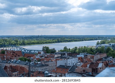 Panoramic elevated view over the rooftops of Torun, Poland, looking toward the wide Vistula river, lush green riverbanks, and the steel Jozef Pilsudski road bridge under a cloudy sky.