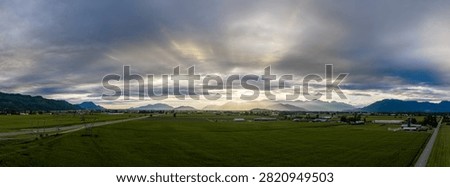 Panoramic evening farmland with dramatic clouds and sun rays over mountains near Chilliwack, British Columbia, Canada.