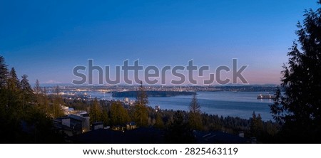 Panoramic high angle view of Vancouver city lights and Lions Gate Bridge across Burrard Inlet during twilight framed by forest foreground.