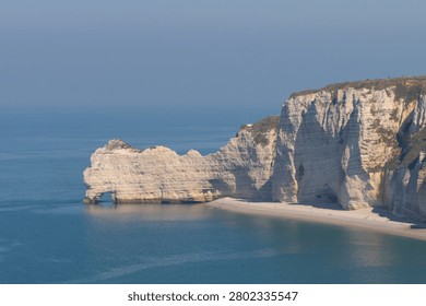A panoramic high-angle view of the iconic white chalk cliffs and the smaller natural stone arch of Étretat, Normandy, France. The massive limestone formations stand out against the calm, misty blue sea and sky on a bright summer day, showcasing the beautiful coastline of the Alabaster Coast.