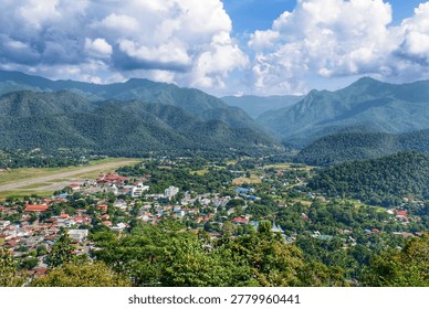 Panoramic high-angle view of Mae Hong Son city in Thailand, showing the airport runway and residential houses surrounded by lush green mountains under a dramatic cloudy sky.