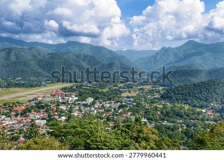 Panoramic high-angle view of Mae Hong Son city in Thailand, showing the airport runway and residential houses surrounded by lush green mountains under a dramatic cloudy sky.
