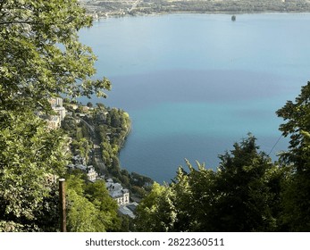 Panoramic high-angle view of a serene blue lake surrounded by majestic mountains, green hills, and a coastal village under a clear sky.