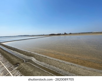 A panoramic, high-angle view of vast salt fields in Thailand, featuring wide shallow pools of seawater separated by earthen dikes and narrow water canals. A rustic wooden storage shed is visible in the background under a bright, cloudless daytime sky, showcasing local coastal industry and agricultural landscape.