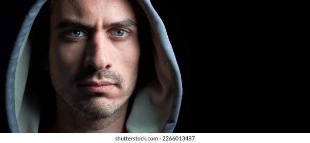 Panoramic horizontal close up portrait of young handsome hooded man with blue eyes looking at the camera with serious tranquil expression isolated on dark black background with copy space. - Powered by Shutterstock