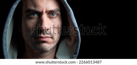 Panoramic horizontal close up portrait of young handsome hooded man with blue eyes looking at the camera with serious tranquil expression isolated on dark black background with copy space.