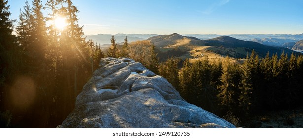 Panoramic image of sunrise over Pysanyi Kamin in Carpathians, Ukraine. Sun's rays pierce through tall pine trees, illuminating rocky foreground. Rolling hills and distant mountain ranges. - Powered by Shutterstock