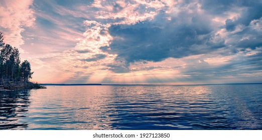 Panoramic Landscape Of Dramatic Sky With Clouds View From The River, The Pink Rays Of The Sun Shine Through The Clouds And Are Reflected In The Ripples Of The Blue Water.