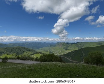 Panoramic landscape featuring rolling green hills and valleys beneath a bright blue sky with scattered fluffy clouds in a mountainous area