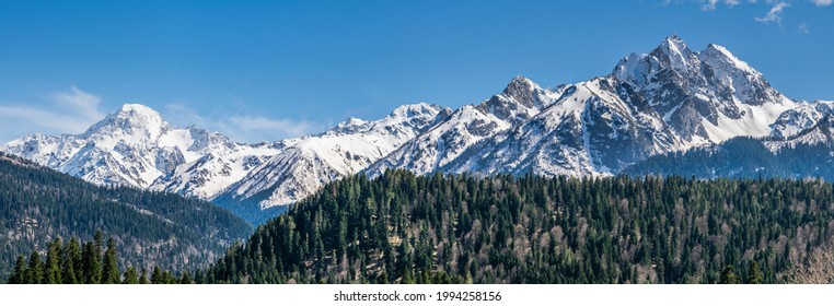Panoramic landscape view, snow covered mountain peaks on mountain range. Hills and mountains covered with green pine forest, Arkhyz villadge, Caucasus mountains, Russia - Shutterstock ID 1994258156