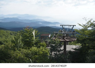 A panoramic mountain view featuring a foreground ski lift mechanism and surrounding lush green trees, overlooking a hazy, layered mountain range under a soft, daylight sky.