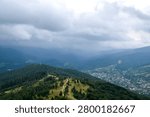 Panoramic mountain view under a dramatic, cloudy sky, featuring rolling green forested ridges and a small village nestled in the valley below. Carpathian Mountains, Ukraine