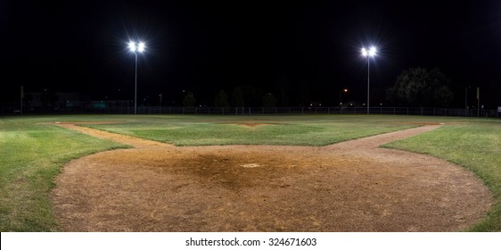 Panoramic night photo of an empty baseball field at night with the lights on taken behind home plate and looking out over the pitcher's mound.