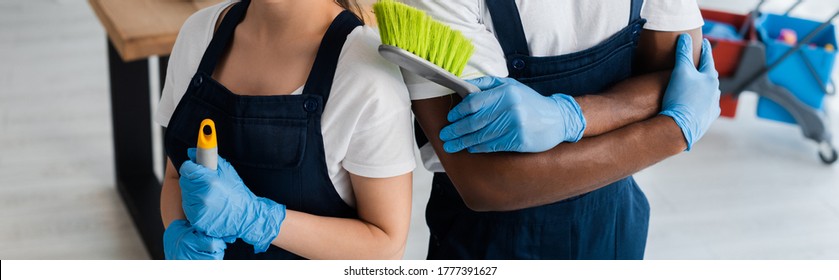 Panoramic orientation of multiethnic cleaners in uniform holding cleaning supplies in office