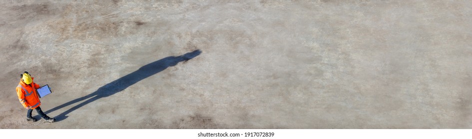 Panoramic Overhead Aerial View Of Anonymous Construction Worker Or Builder Wearing A Hi Vis Jacket, A Hardhat And Carrying A Clipboard On A Building Site With Their Shadow Panorama Web Banner Header