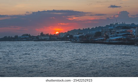 A panoramic photo of a coastal city at sunset. The image features a vibrant orange and pink sky, calm water, and a cityscape with buildings and structures silhouetted against the colorful backdrop. - Powered by Shutterstock