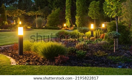 Panoramic Photo of LED Light Posts Illuminated Backyard Garden During Night Hours. Modern Backyard Outdoor Lighting Systems.