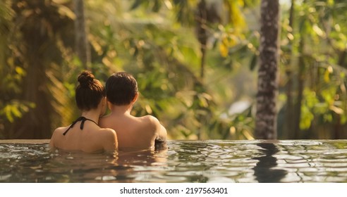 Panoramic photo of young adult couple in love swimming together in open-air infinity pool enjoying tropical vacation on villa looking away at rainforest on sunset. High quality photo - Powered by Shutterstock