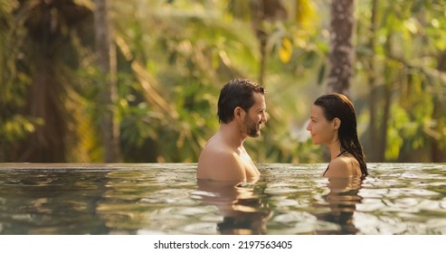 Panoramic photo of young adult couple in love swimming together in open-air infinity pool enjoying tropical vacation on villa looking to each other against rainforest on sunset. High quality photo - Powered by Shutterstock