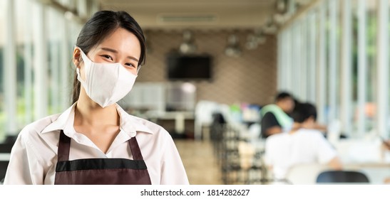 Panoramic Portrait Attractive Asian Waitress Wear Face With Background Of Customers Sit In Social Distance Table In Restaurant Indoor. New Normal Restaurant Lifestyle Concept.