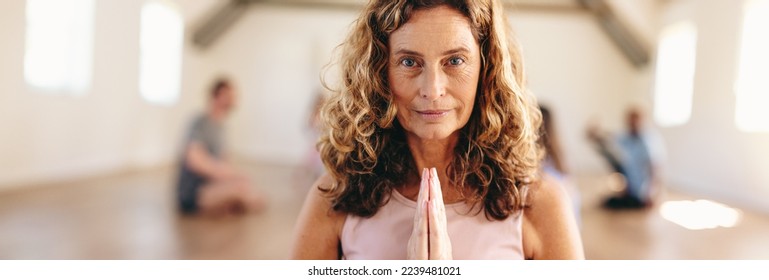 Panoramic portrait of a mature woman meditating in prayer pose with a group of people in the background. Senior woman practicing yoga with her class in a community fitness studio. - Powered by Shutterstock