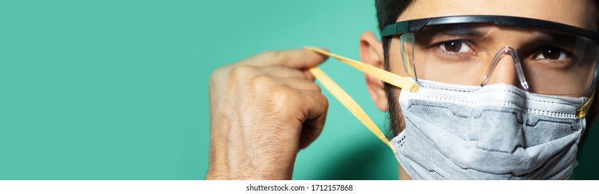 Panoramic Portrait Of Young Guy Putting On Medical Face Mask, Wearing Safety Goggles Against Coronavirus On Background Of Aqua Menthe Color With Copy Paste.