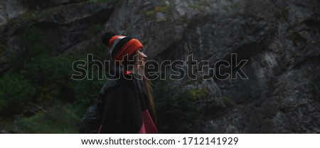 Panoramic portrait of young traveler girl in mountains on background of stone rock. Wearing scarf and orange hat.