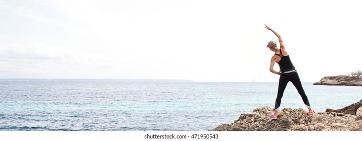 Panoramic Rear View Of A Healthy Mature Woman Figure Stretching And Exercising On A Rock Platform By Blue Sea In A Nature Coastal Destination, Outdoors. Fitness And Body Care Senior Lifestyle.