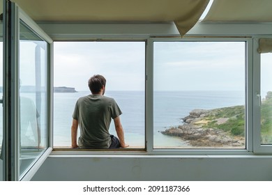 Panoramic rear view of unrecognizable man sit on a window frame looking at the sea. Horizontal view of man isolated at home with a view from the window of the blue ocean on the background. - Powered by Shutterstock