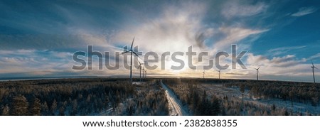 Panoramic scenic aerial photo over forest winter road with windmills standing in row at the left side in forest. Road in the middle of view. Large wind turbines with blades. High altitude