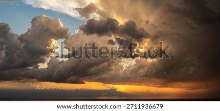 Panoramic seascape view of dramatic stormy sky with dark cumulonimbus clouds and bright orange sunlight beams over the calm water horizon during a beautiful sunset evening on the sea coast.