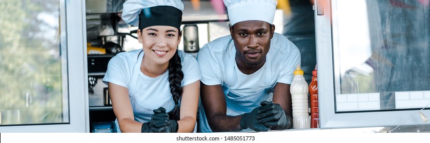 Panoramic Shot Of Asian Girl And African American Man In Hats Smiling From Food Truck 