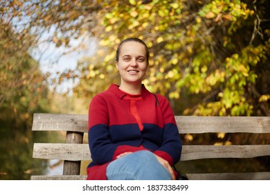 Panoramic Shot Of Beautiful Girl In Casual Clothes With No Make Up Sitting On Wooden Bench In Park Near Lake, Looks Smiling At Camera, Female Having Rest On River Bank.