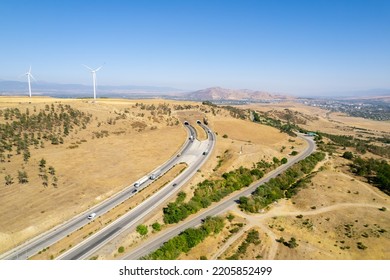 Panoramic Shot From A Bird's Eye View Of Wind Turbines In The Middle Of Yellow Fields And A Road With A Tunnel Among The Mountains. Wind Turbine Construction Against The Backdrop Of Mountains To Gener