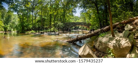 A panoramic shot of Boulder Bridge in Rock Creek Park in Washington, DC on a summer day. Rock Creek flows underneath the iconic bridge; a fallen tree is seen in the foreground.