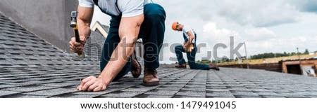 panoramic shot of handyman holding hammer while repairing roof near coworker 