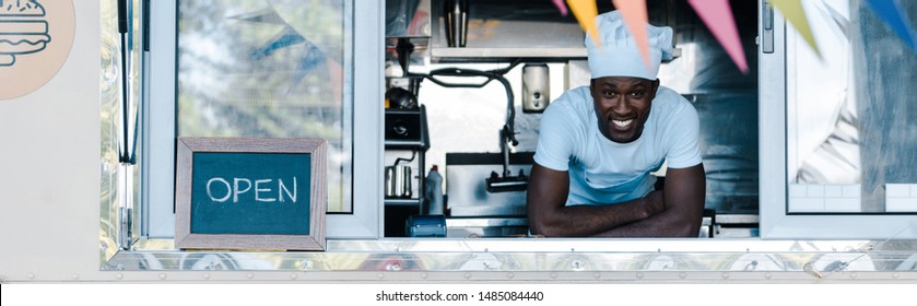 Panoramic Shot Of Happy African American Man In Chef Uniform Smiling From Food Truck 