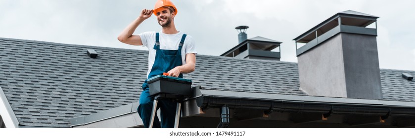 Panoramic Shot Of Happy Repairman Sitting On Roof And Holding Toolbox 