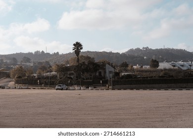 A panoramic shot of a large, empty, unpaved parking lot (parking) with a low divider wall, set against a background of low houses, green foliage, and hills under a bright, cloudy sky.