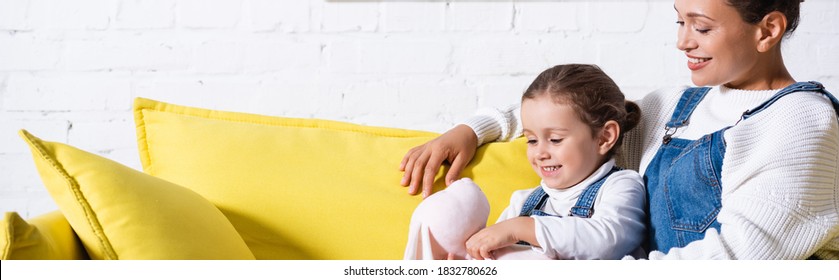 Panoramic Shot Of Mom And Daughter With Soft Toy Sitting On Yellow Couch At Home