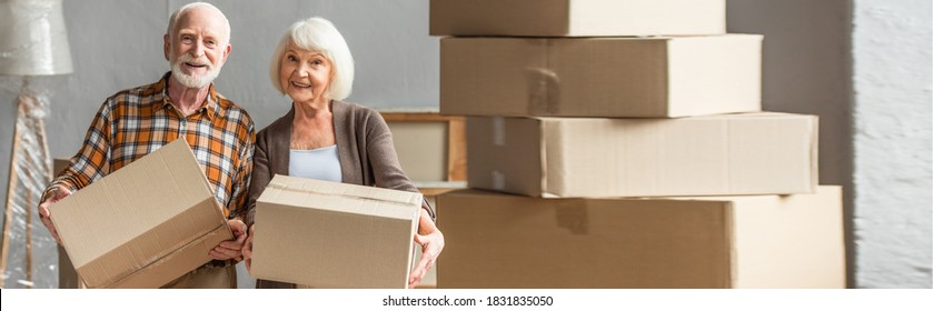 Panoramic Shot Of Senior Couple Holding Cardboard Box And Looking At Camera In New House, Moving Concept