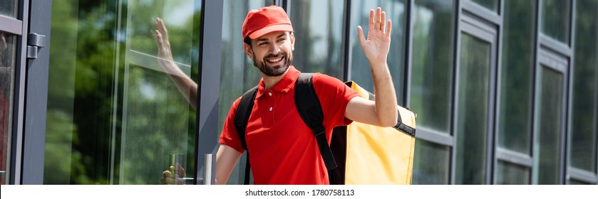 Panoramic Shot Of Smiling Delivery Man Waving Hand Near Building On Urban Street