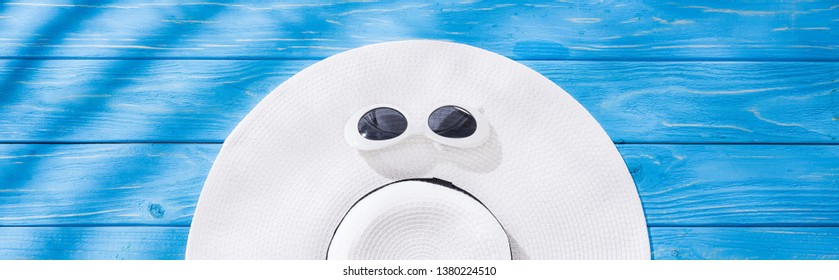 panoramic shot of white floppy hat with black ribbon and sunglasses on blue wooden background