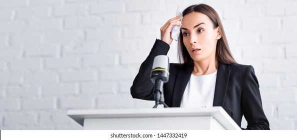 panoramic shot of worried lecturer suffering from fear of public speaking holding napkin near forehead while standing on podium tribune - Shutterstock ID 1447332419