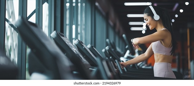 Panoramic Side view of beautiful young asian woman running on treadmill and listening to music via headphone with arm a smart watch for tracking speed during sports training in a gym. - Shutterstock ID 2194326979